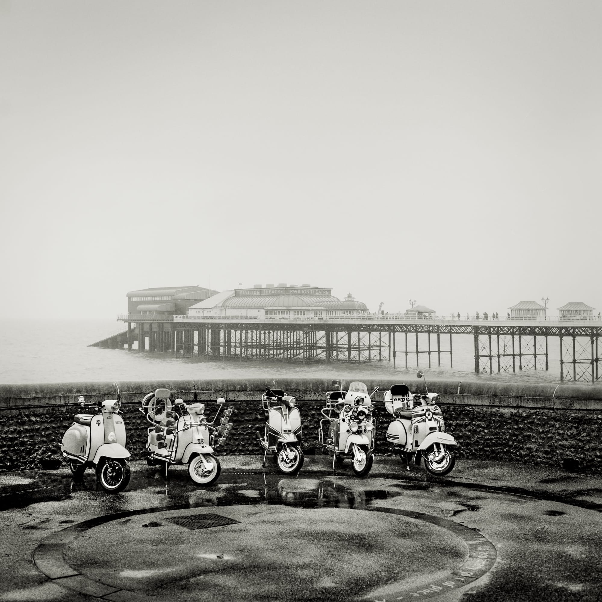 Row of vintage scooters parked on a wet pavement, with a pier and theatre in the background.