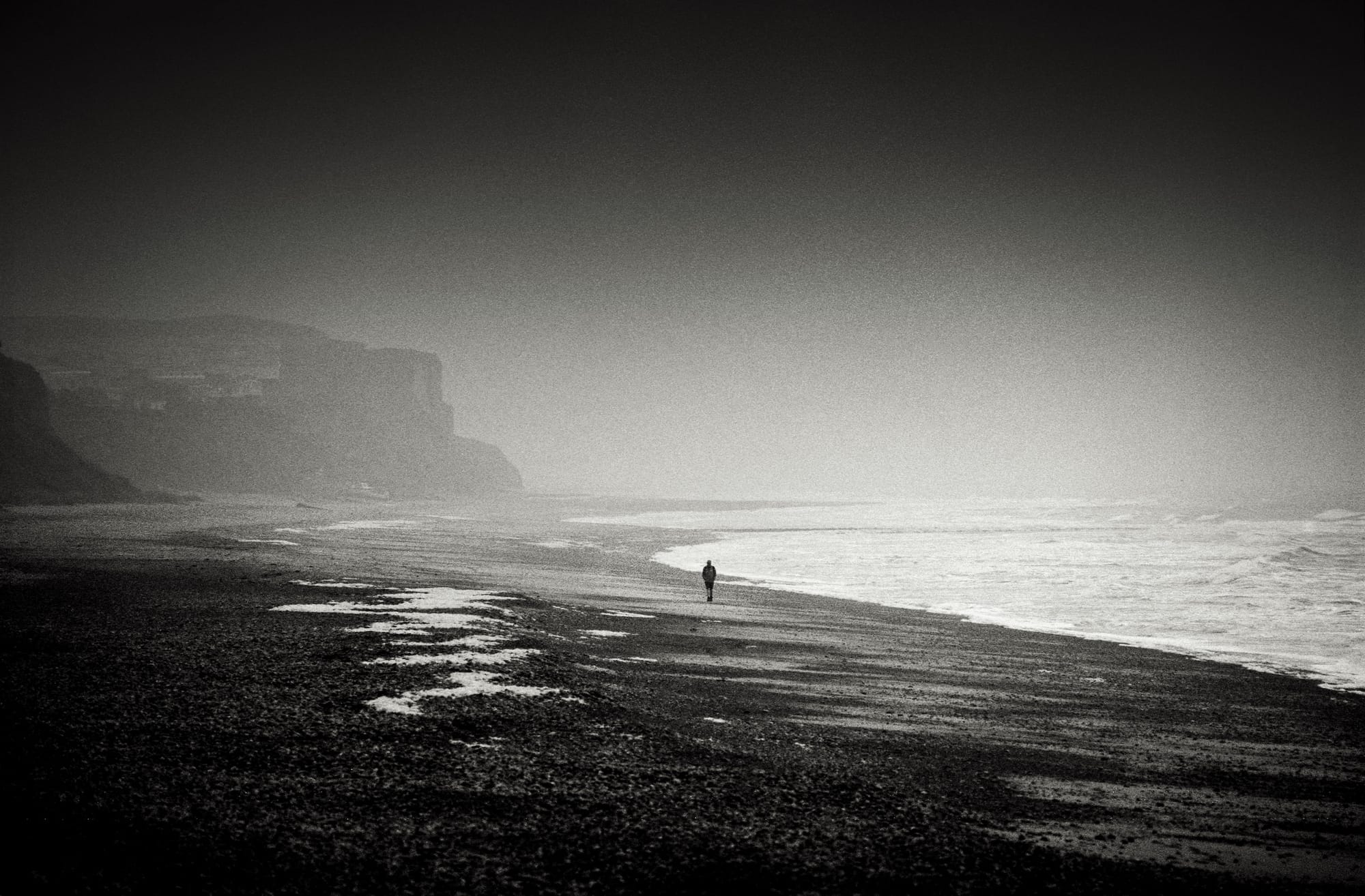 Foggy seaside scene with a lone person strolling along the wet sand, ocean waves in the background.