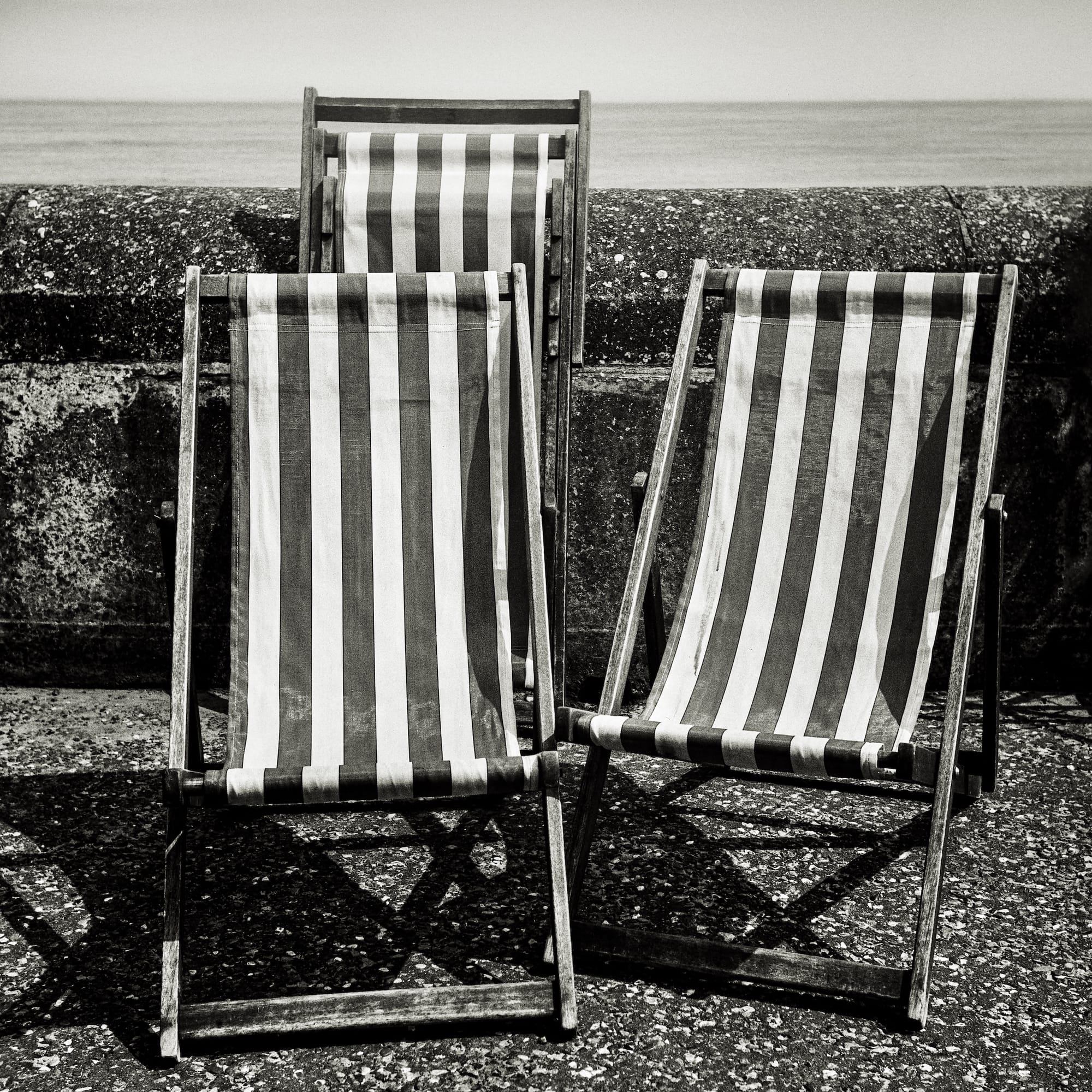 Three vintage deck chairs with striped fabric, positioned on a stone promenade, overlooking the ocean.