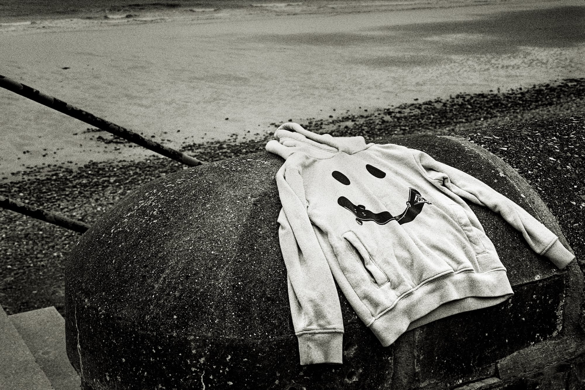Black and white photo of a hoodie with a playful face design, laid on a stone wall near the sea.