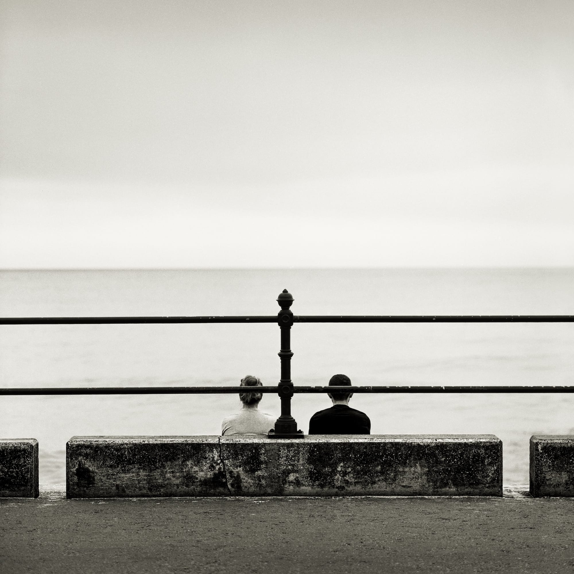 Black and white photo of two people sitting on a stone wall overlooking the sea.