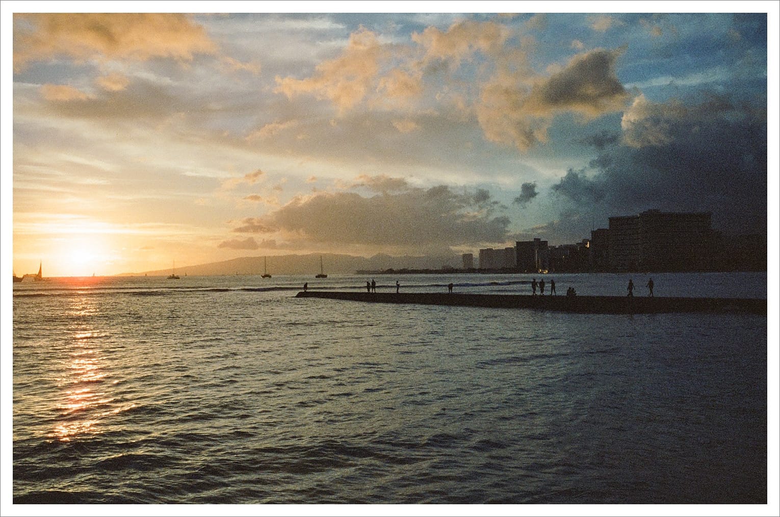 Evening scene with sunset, people on a pier and buildings in the distance.