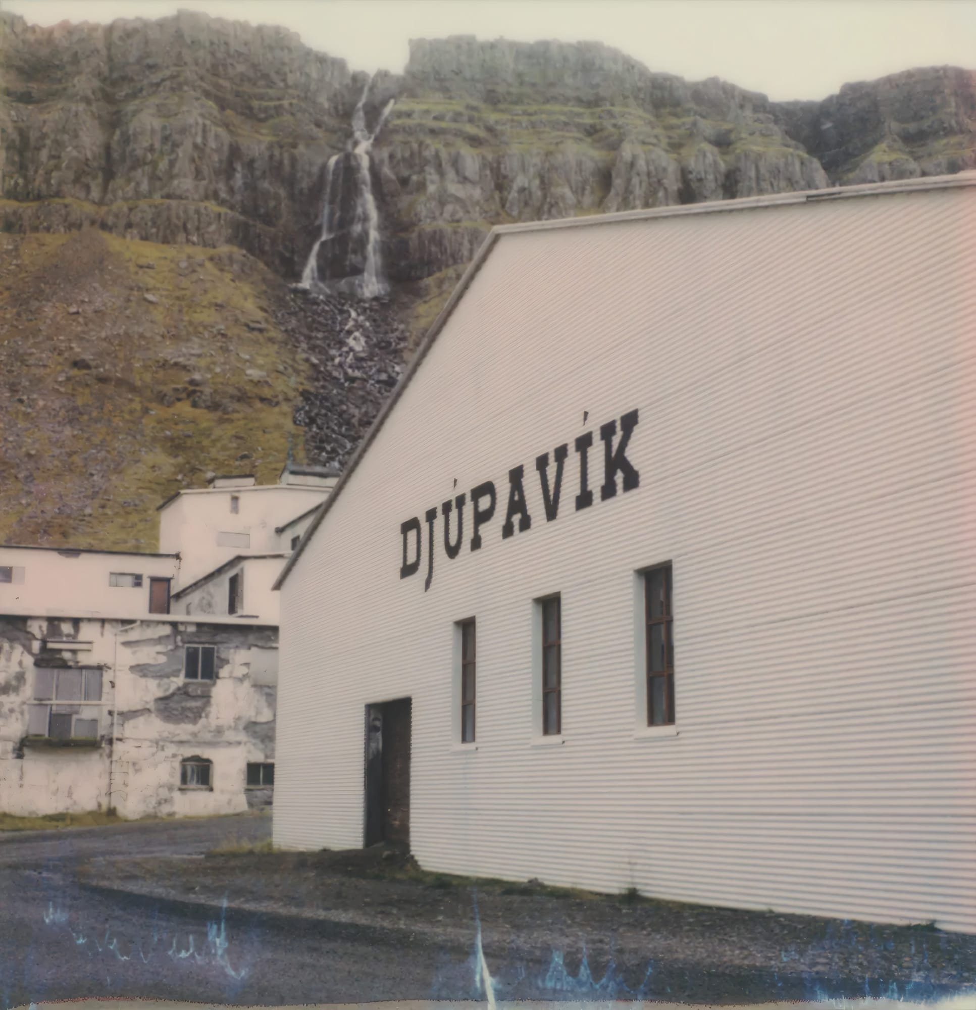 Building with "DJÚPAVÍK" sign and waterfall behind cliffs on Polaroid.