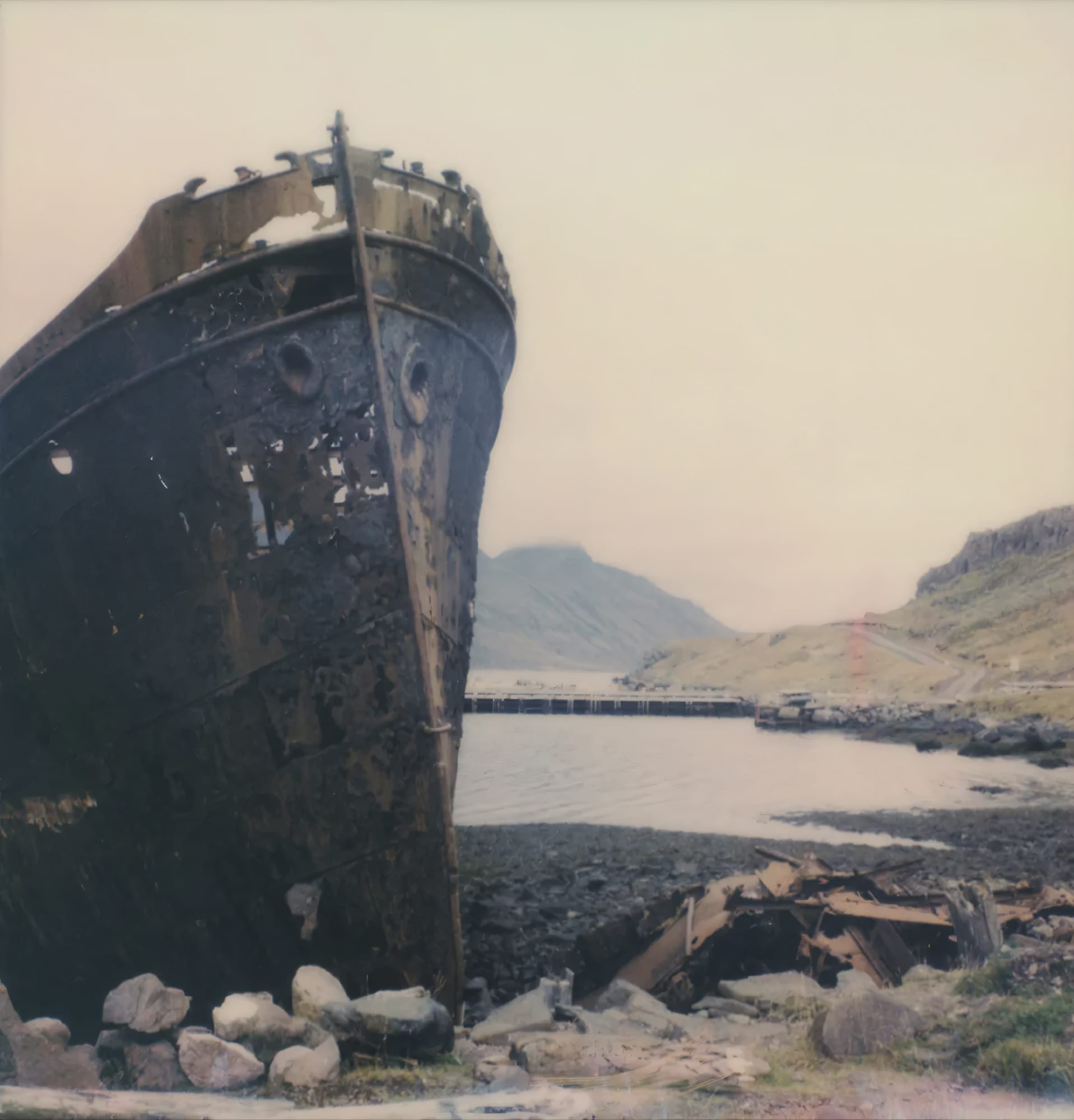Rusted shipwreck bow on rocky shore with mountains behind on Polaroid.