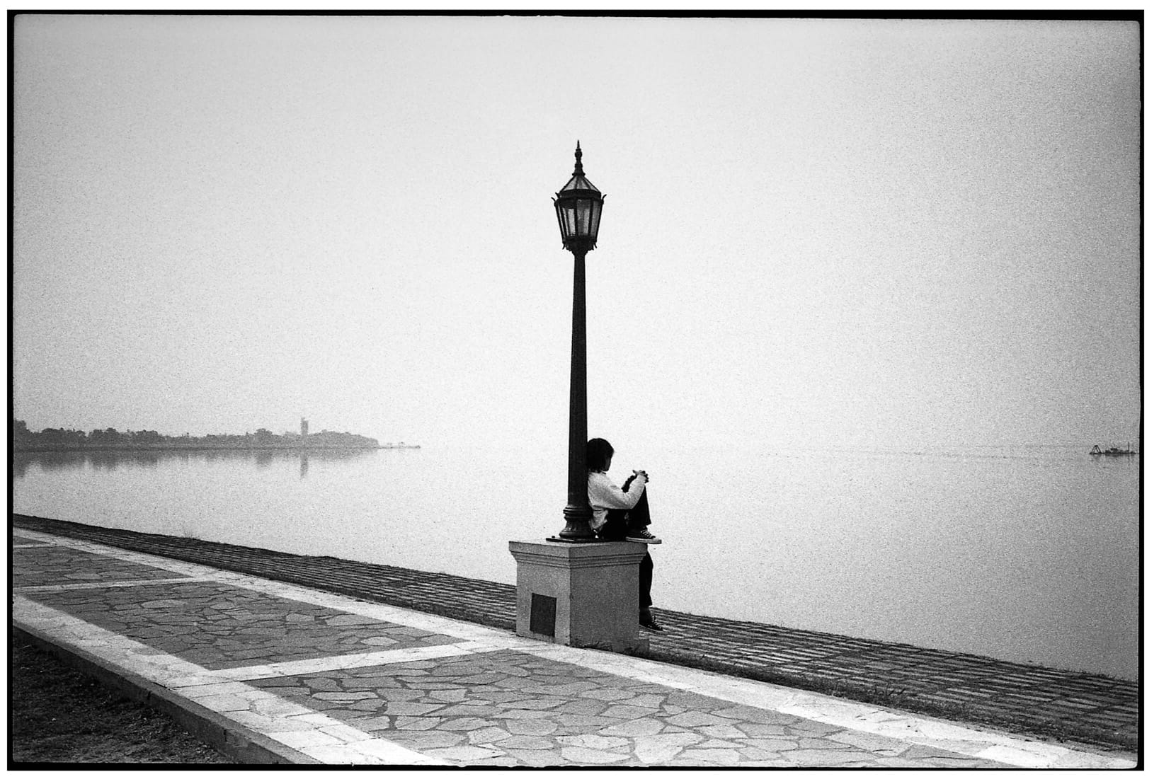 Person sitting by lamp post on waterfront promenade.