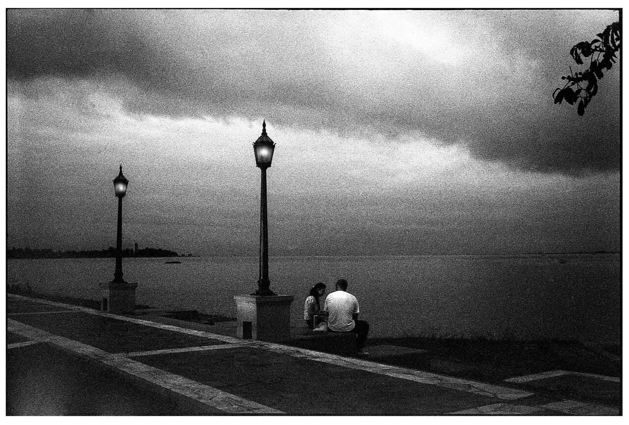 Two people sitting by lamp posts at waterfront at dusk.