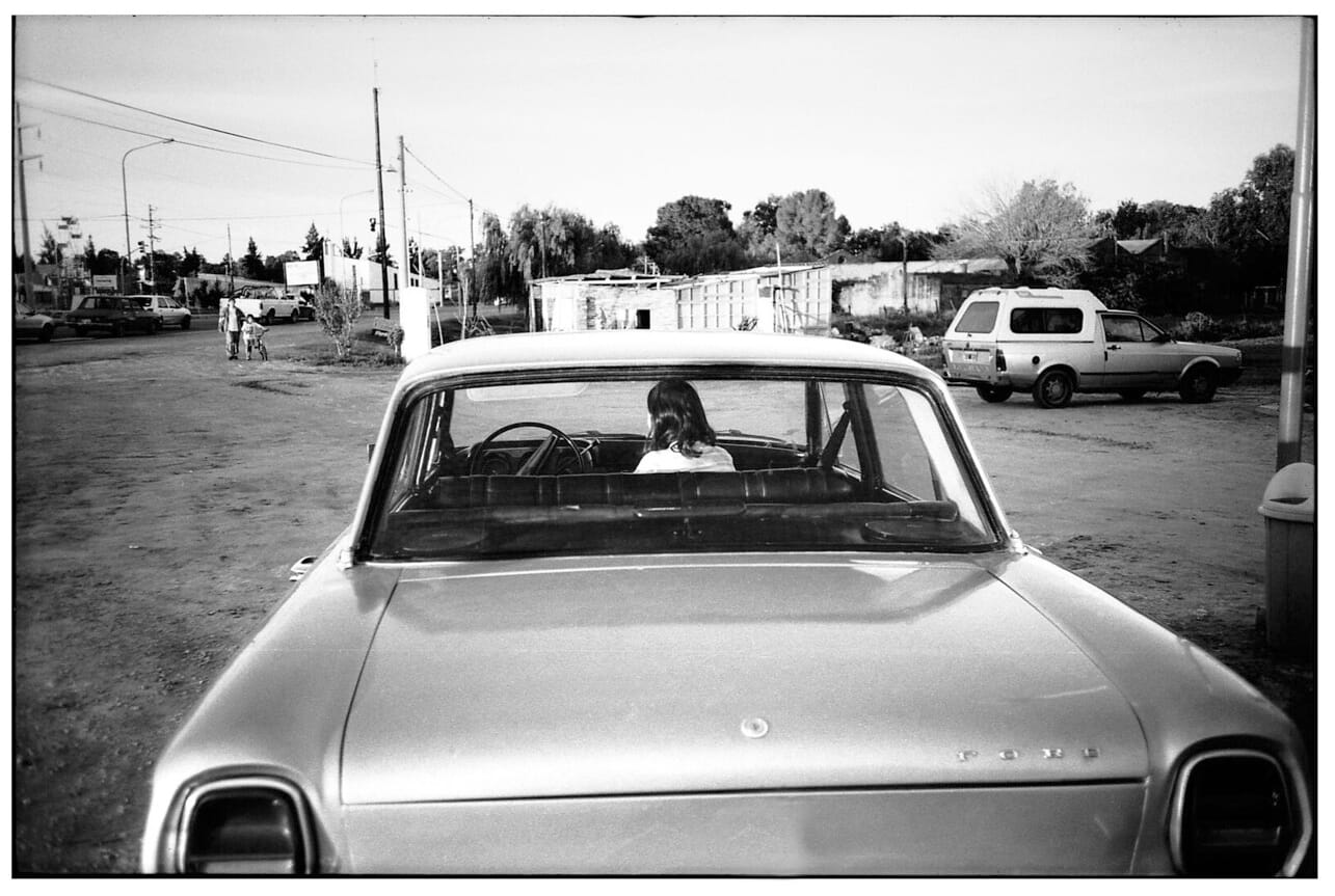 Young woman in back seat of vintage Ford in dirt parking lot.