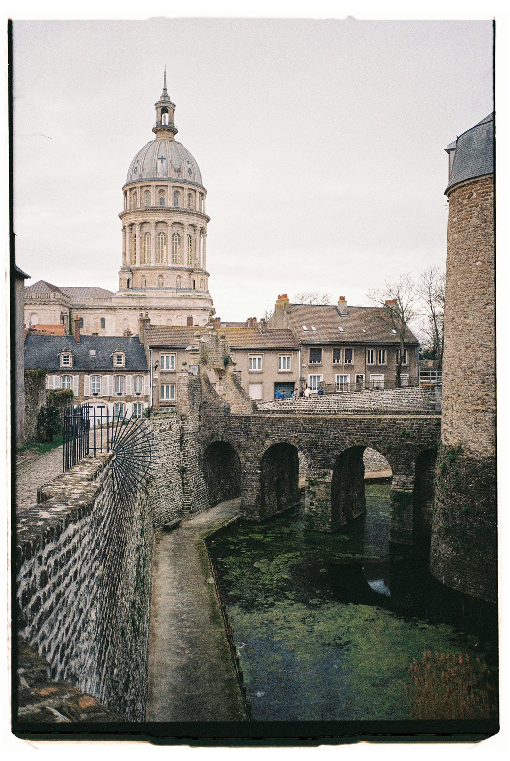 Stone aqueduct with arches over green water, medieval basilica dome rising behind row houses.