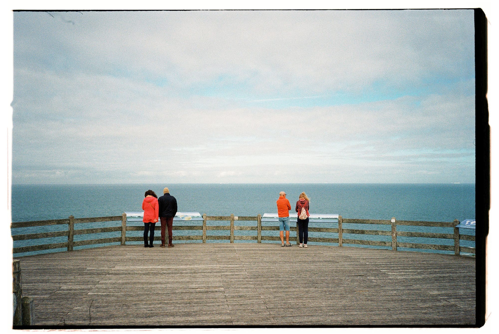 Four people standing at wooden observation deck railing facing ocean horizon under cloudy sky.