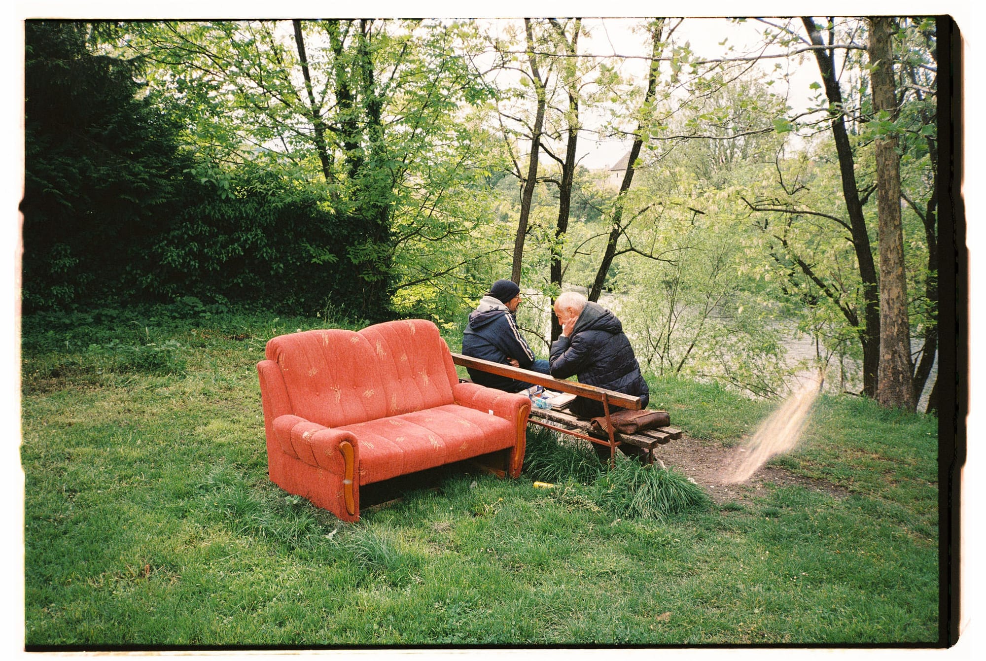 Orange vintage sofa on grass hillside, two people seated on wooden bench, dirt path and forest beyond.