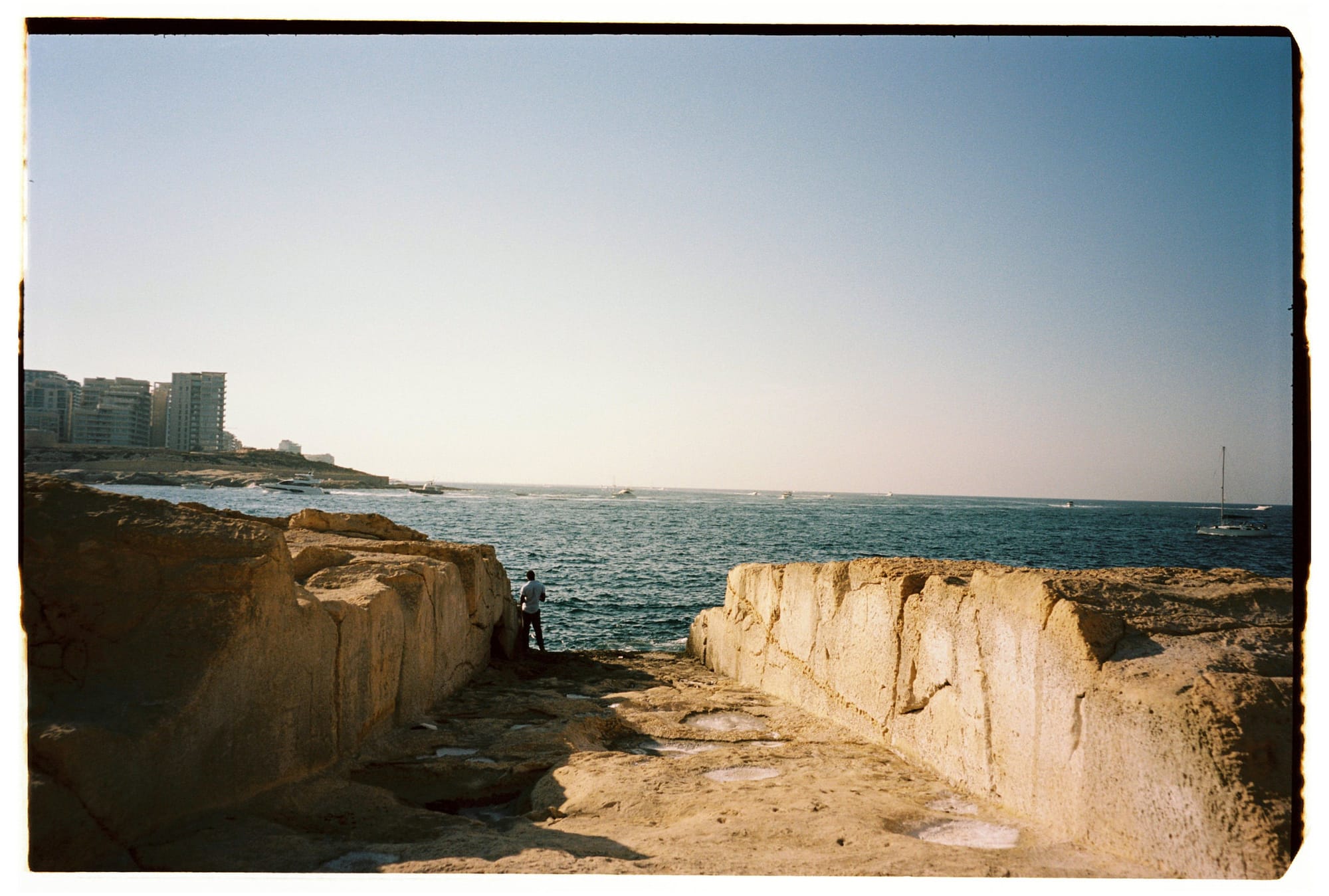 Rocky coastal cliff formation, figure standing on sandstone ledge, Mediterranean sea and distant buildings.