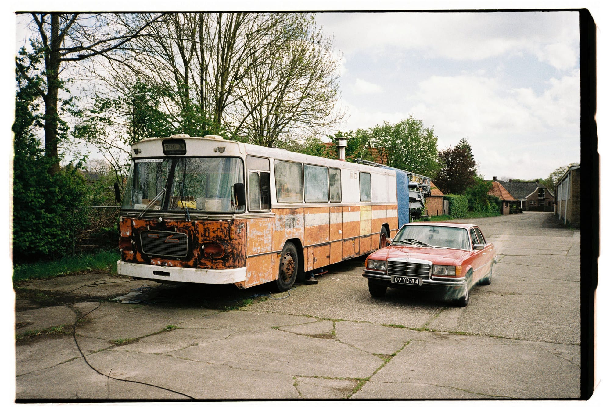 Rusted orange and white bus beside red Mercedes sedan with visible license plate in cracked asphalt lot.