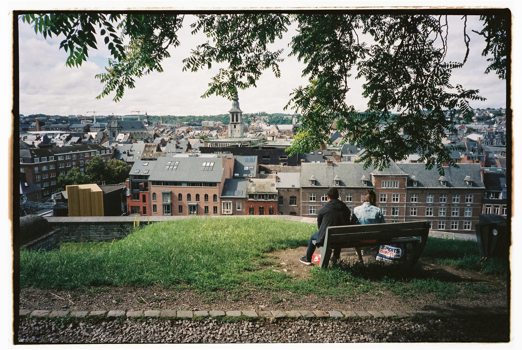 Two figures on wooden bench under tree canopy overlooking dense European city rooftops and church spire.
