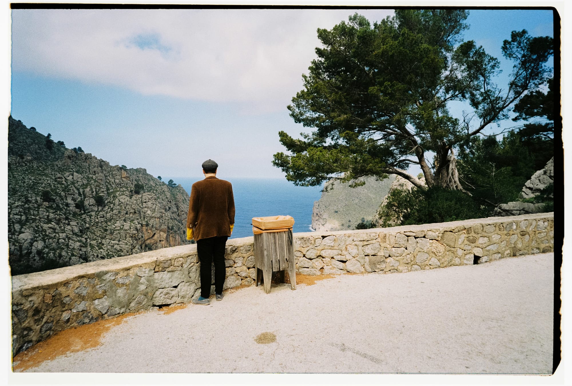 Man in brown jacket and flat cap facing coastal cliff vista, wooden trash bin and stone wall.