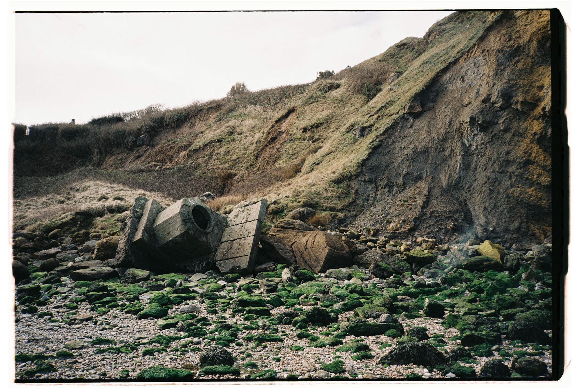 Concrete bunker remnants with circular opening on rocky beach, eroded cliff face and green algae.
