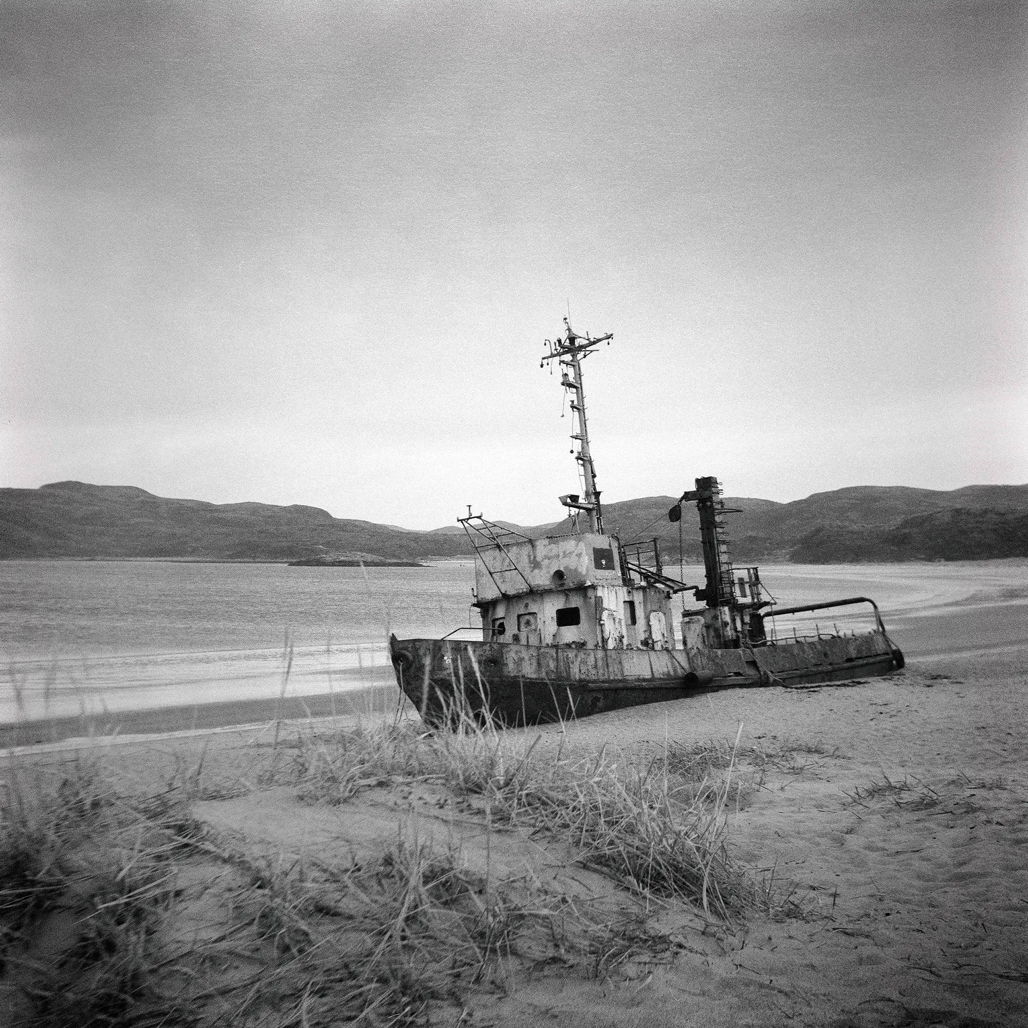 Abandoned weathered ship tilted on sandy beach with mountains across water in distance.