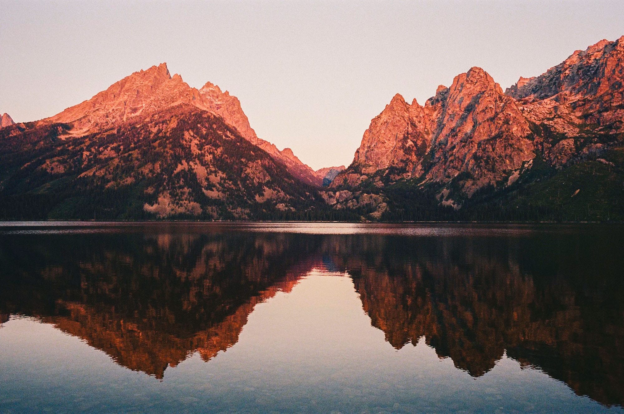 Jagged peaks reflect in still lake under orange sunrise light.