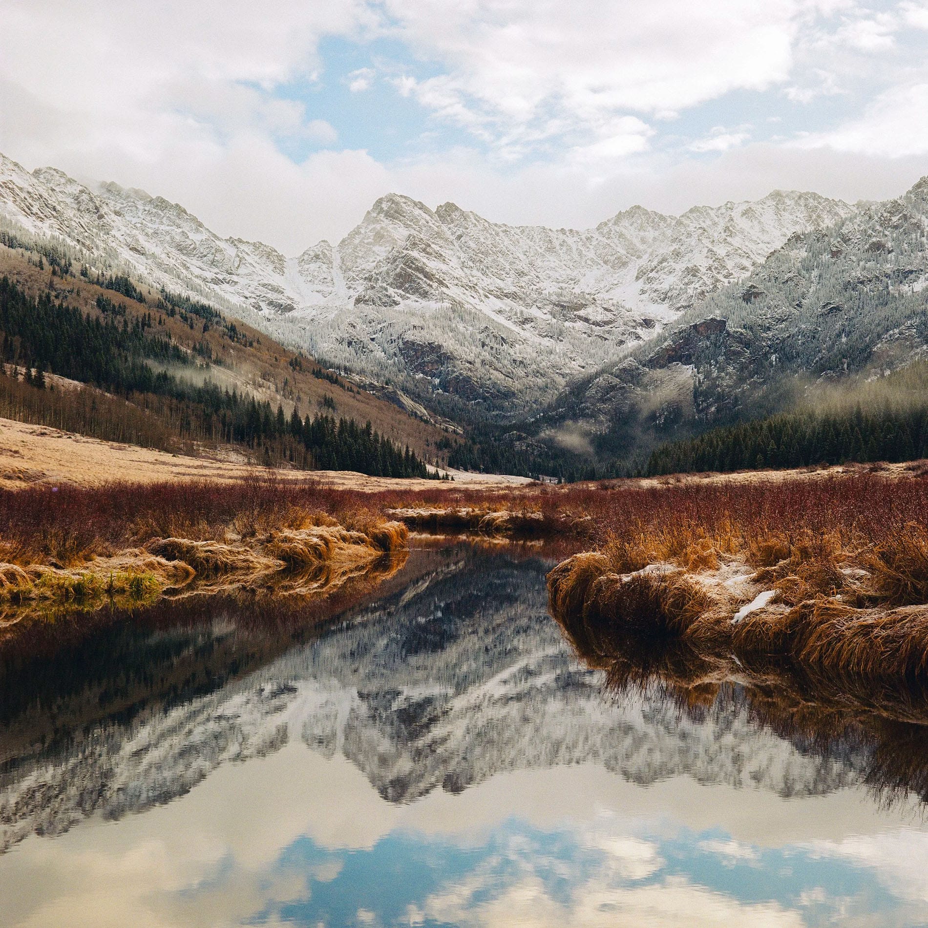 Snow-dusted mountains reflect perfectly in stream with golden grasses.