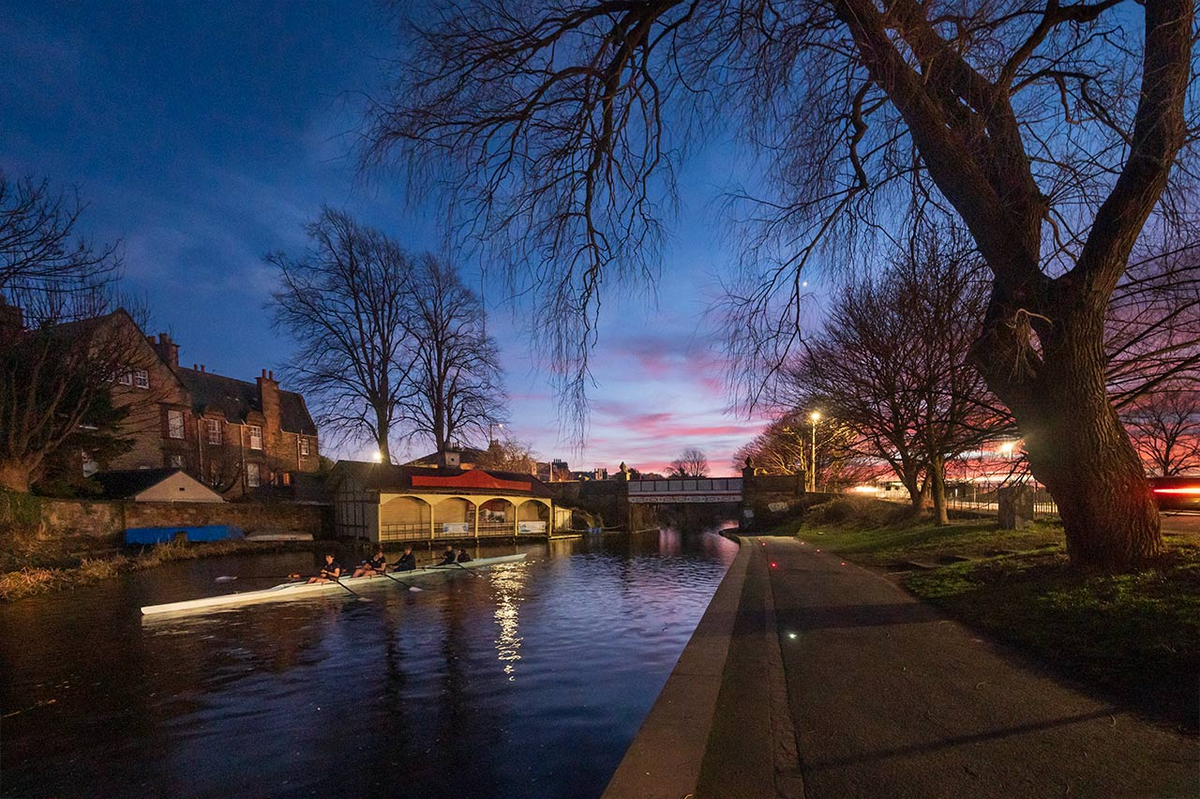 People in a rowing boat on a canal flanked by trees with a sunset in the background. 