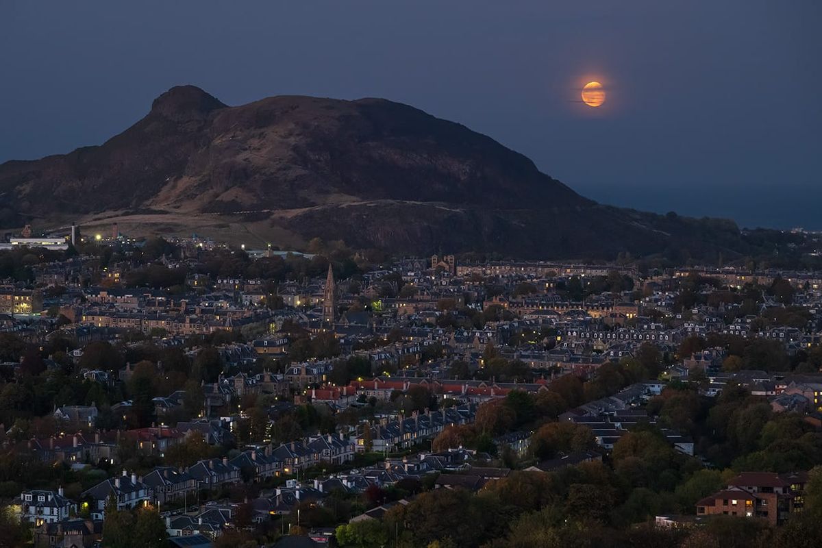A moon rising next to a hill behind a city skyline. 