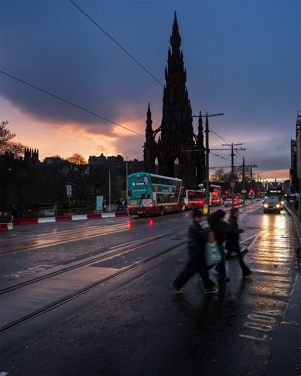 People crossing a city street with traffic and buildings in the background. 