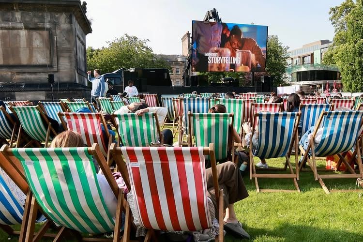 Rows of people sitting in deck chairs watching a large screen in a city park. 