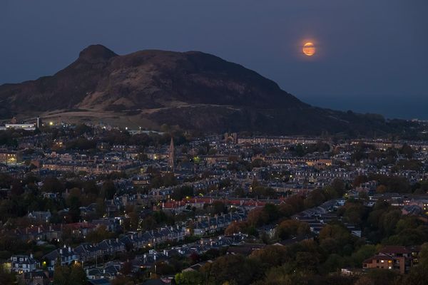 A moon rising next to a hill behind a city skyline. 