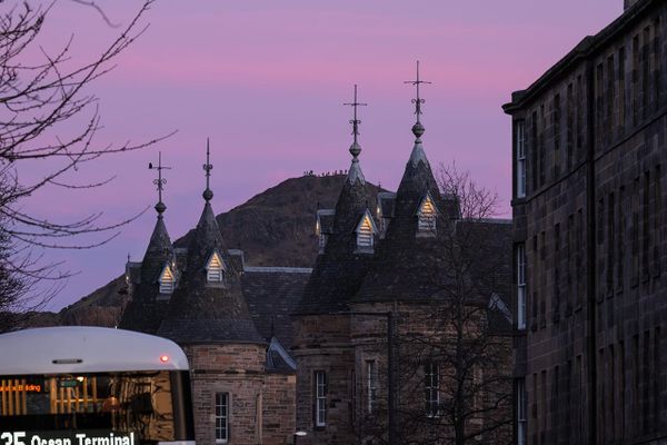 Silhouettes of building roofs against a pink sky with a hill in the background and a bus in the foreground. 