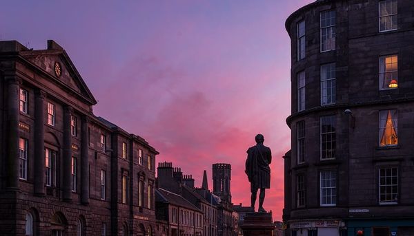 The silhouette of a statue against a pink and purple sky with buildings either side. 