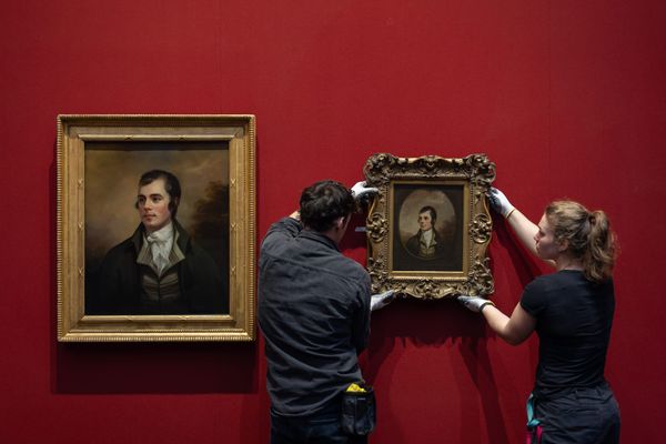 Two people hanging a framed painting on a red wall next to a similar but larger portrait. 