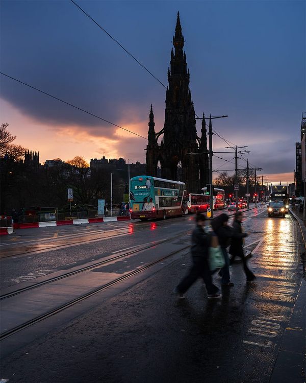 People crossing a city street with traffic and buildings in the background. 