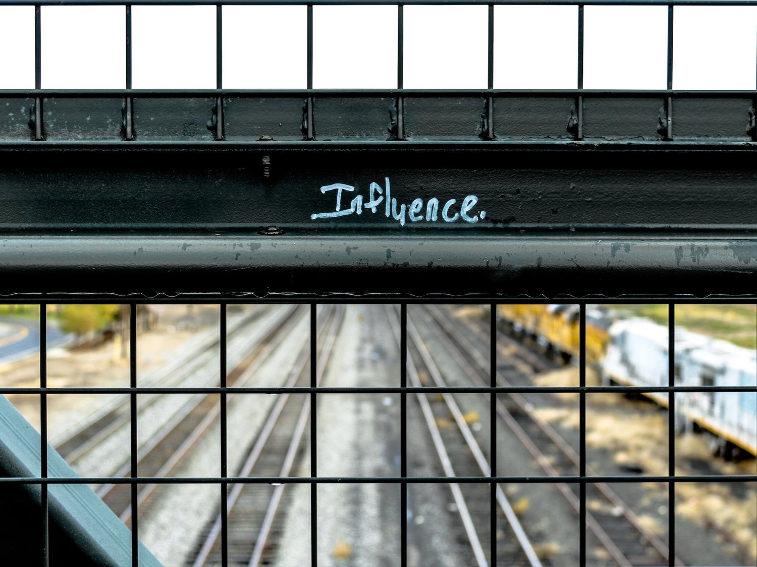 The word "Influence" is inscribed in white marker on a black wired fence that spans a railway track.