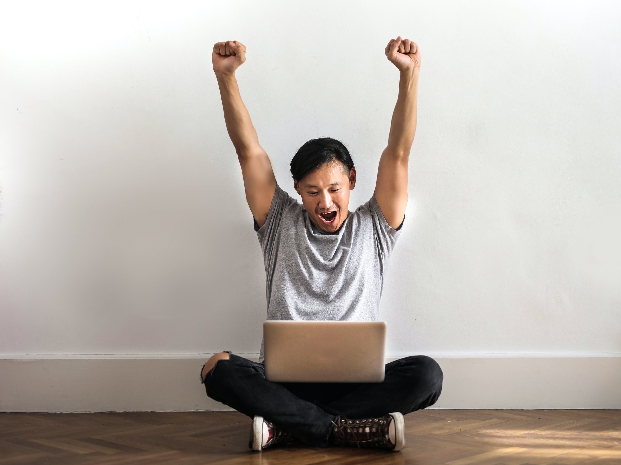 A man in a grey t-shirt and black trousers sits on a wooden floor and works on his laptop, his hands in the air in celebration.