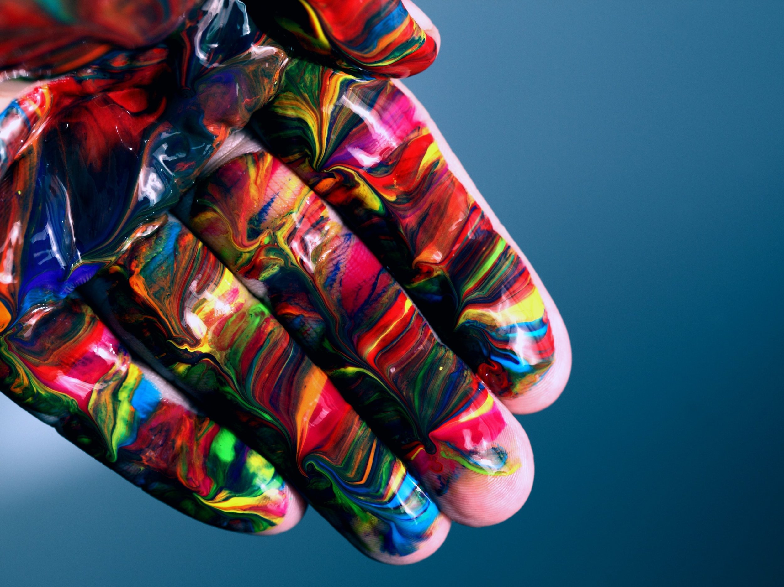 An open hand covered with vibrant, multi-colored paint, displaying a blend of red, blue, yellow, and green hues with swirls and marbling effects against a blurred blue background.