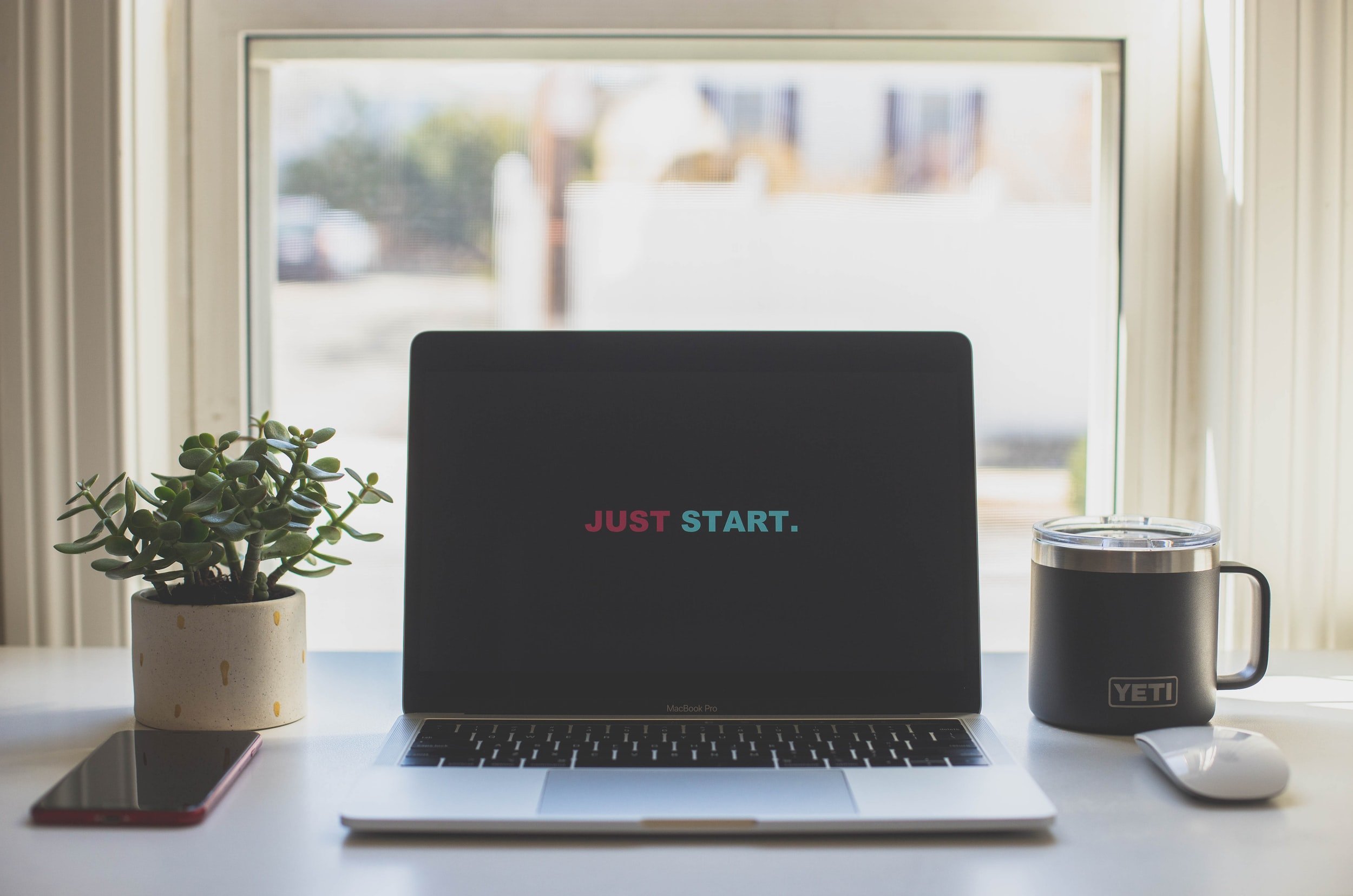 An Apple laptop in front of a window with a Yeti mug, an iPhone, and a magic mouse with the words "Just Start" on the screen&nbsp;