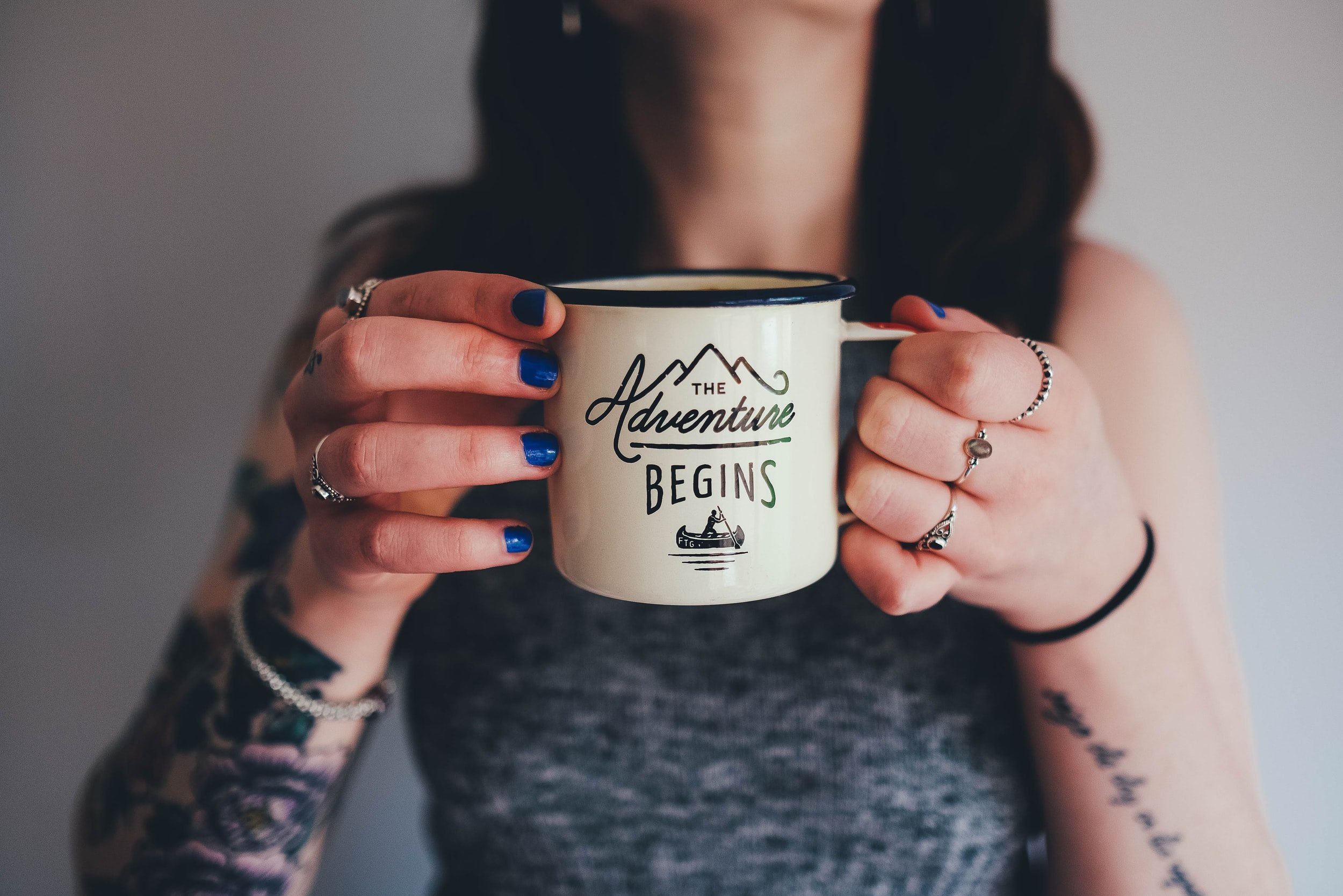 A tattooed woman holds an enamel mug emblazoned with the words "The adventure begins."