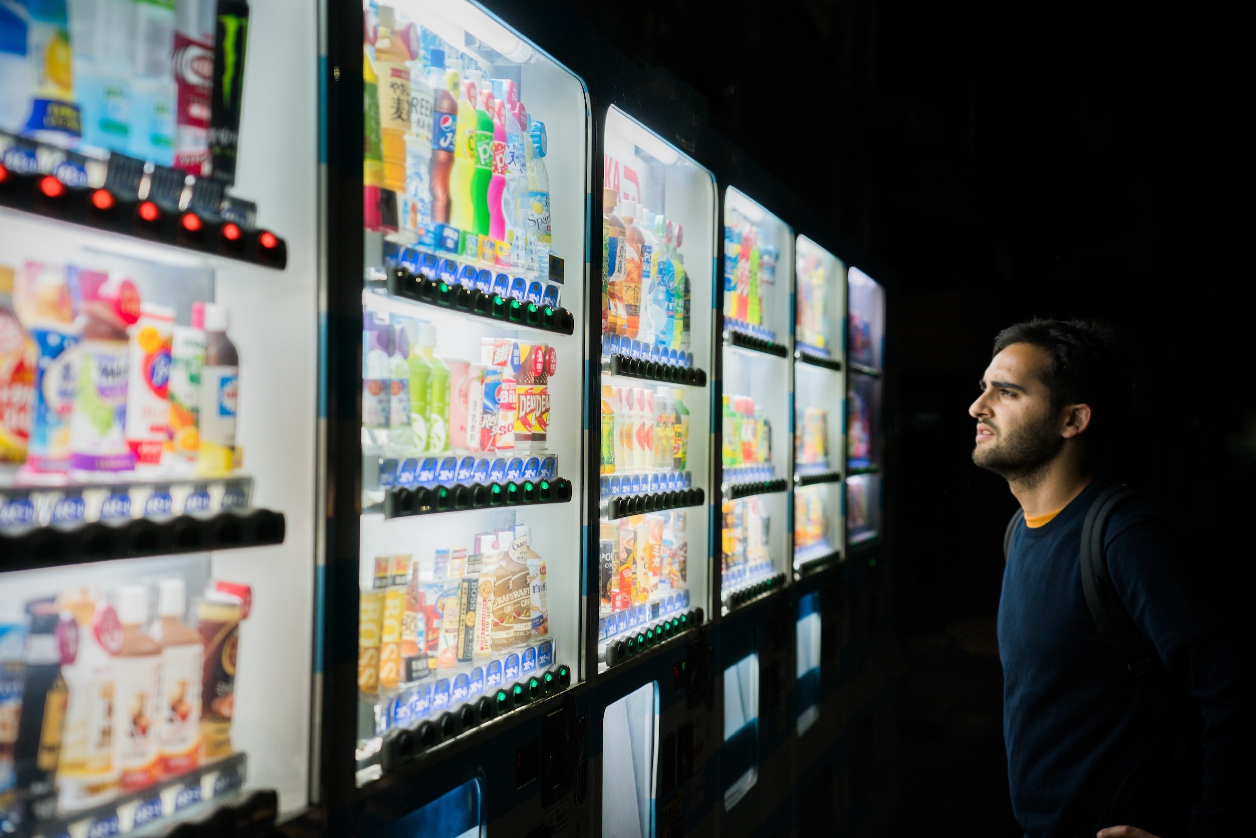 A man pondering which drink to select from a plethora of beverage vending machines.