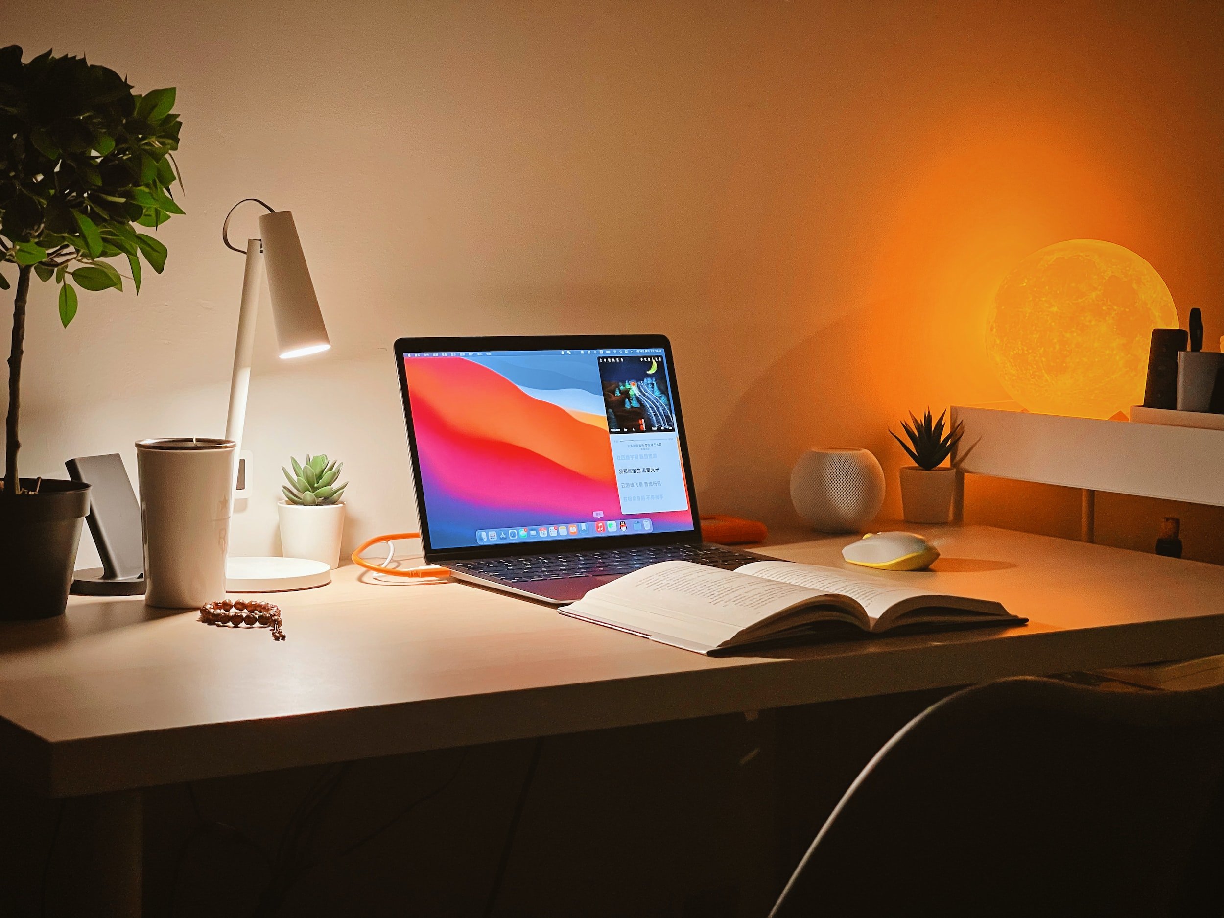 On a desk with a lamp, an open Apple laptop rests next to an open book.