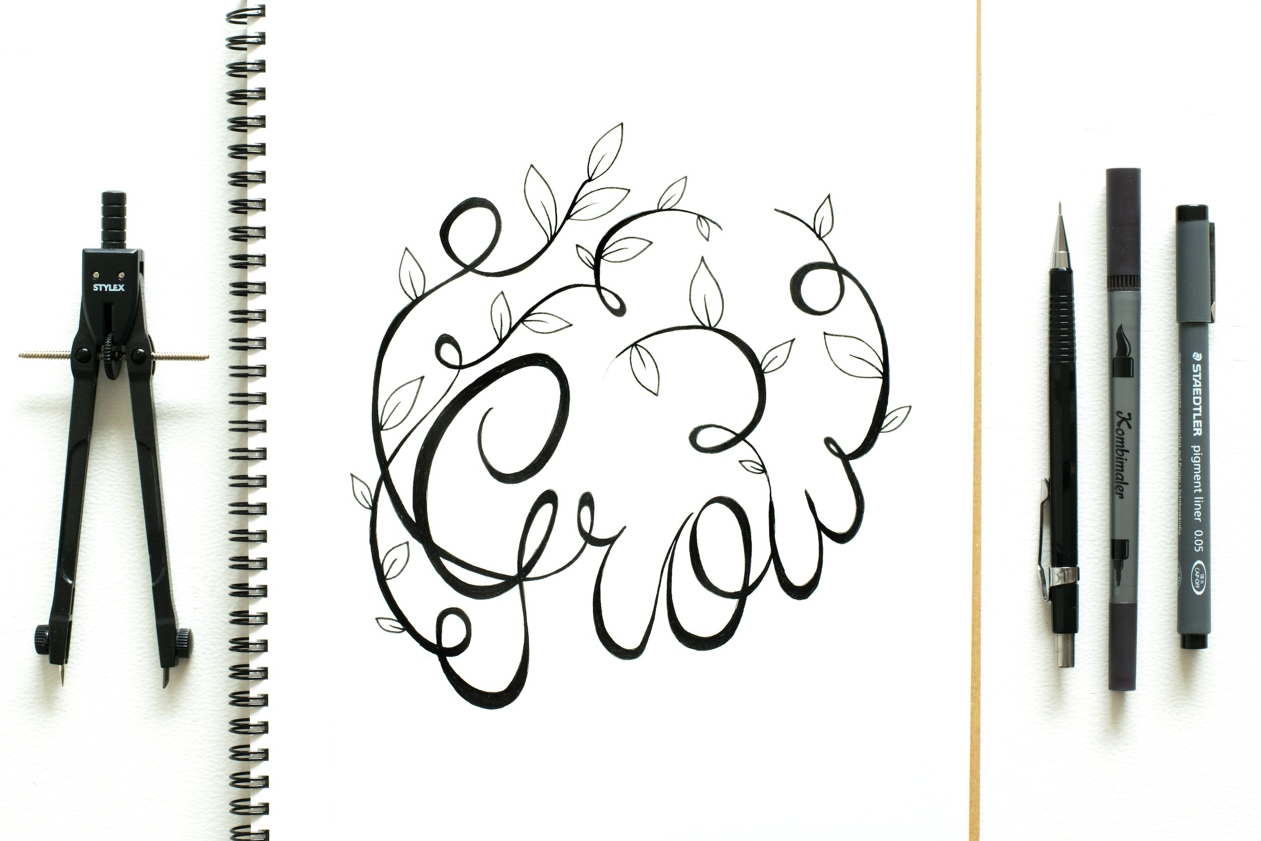 The word "Grow" is hand-lettered in black ink with leaves on a sketch pad alongside a mechanical pencil and ink pens.
