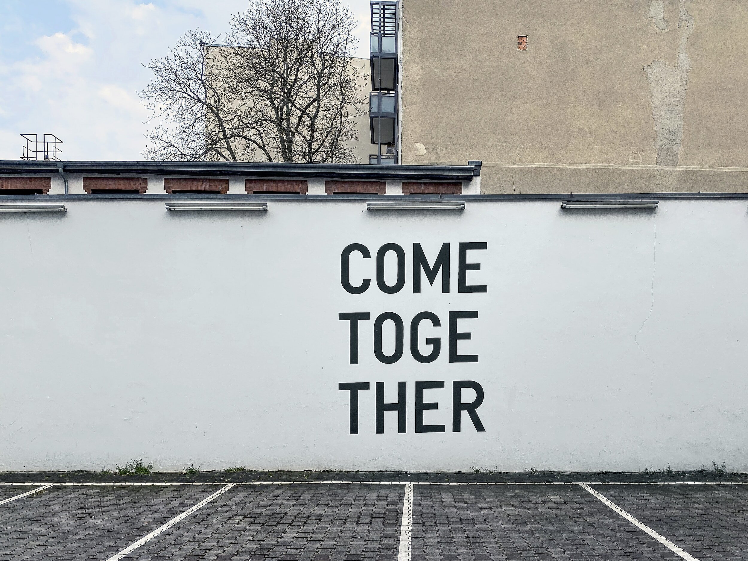 On a large white wall in a car park, the words "Come Together" are painted in black.
