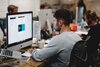 A man works on a graphic design project on an iMac in a busy office, with colleagues in the background and various work tools on his desk.
