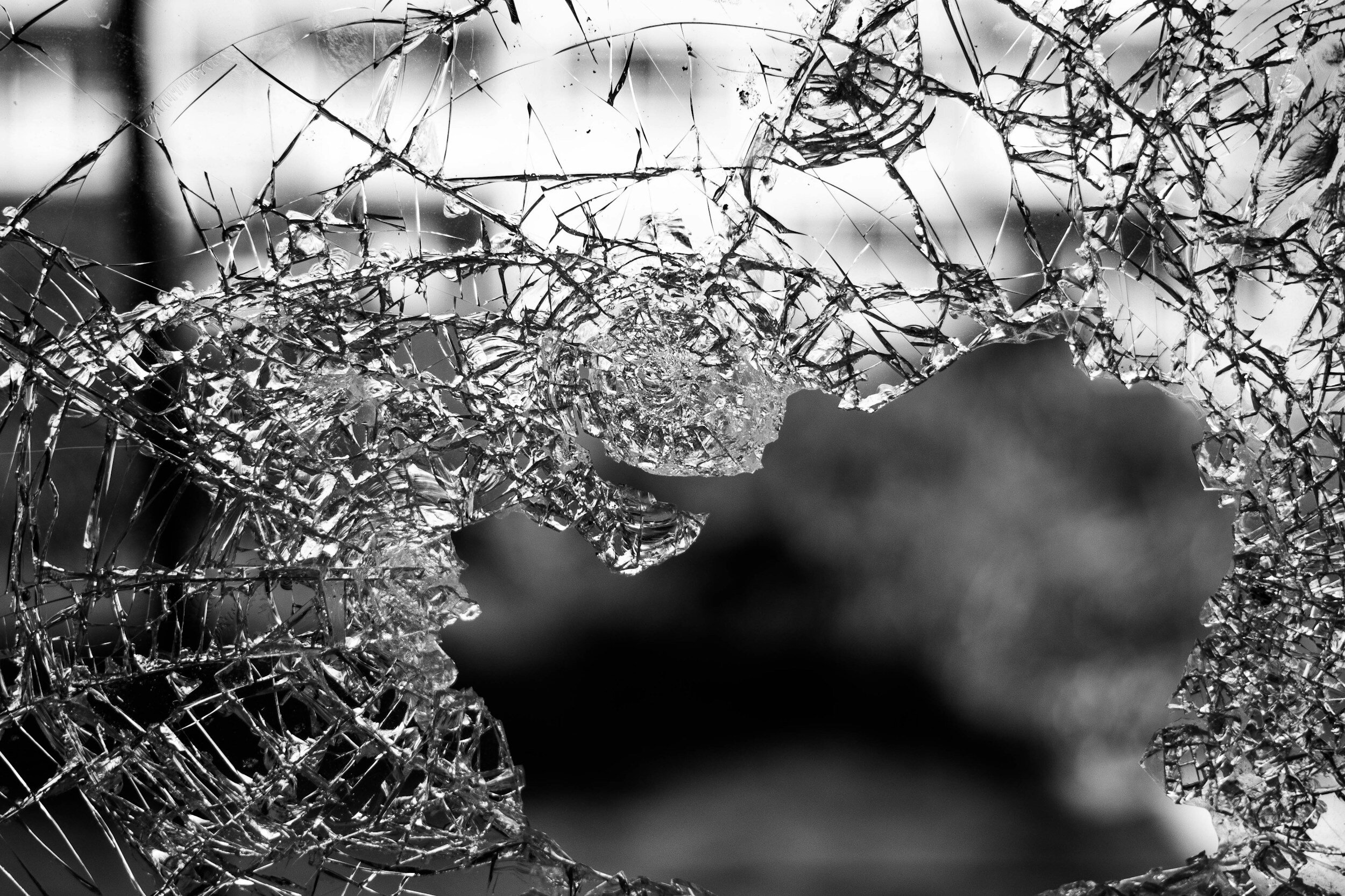 A black and white photo of a shattered glass pane, with cracks creating a web-like pattern and gaps revealing a blurred background.