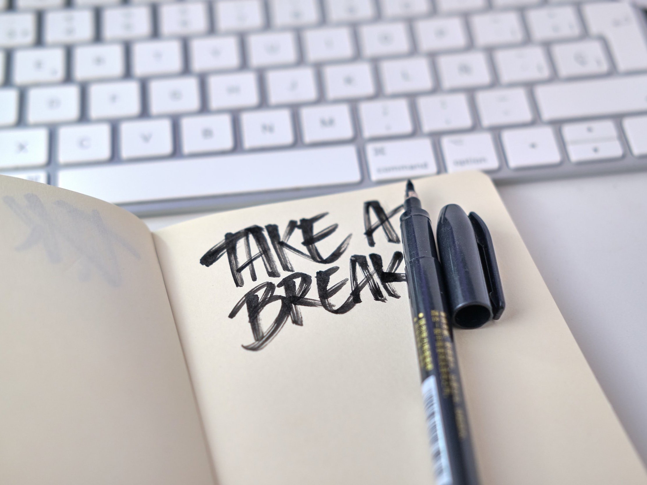 "Take A Break" is handwritten in a notepad with a marker in front of an Apple keyboard.