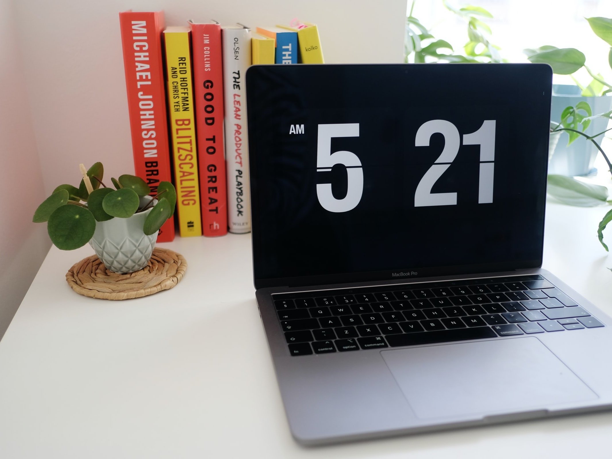 An Apple laptop in front of a selection of productivity books displays the time at 5:21 am on the screen.