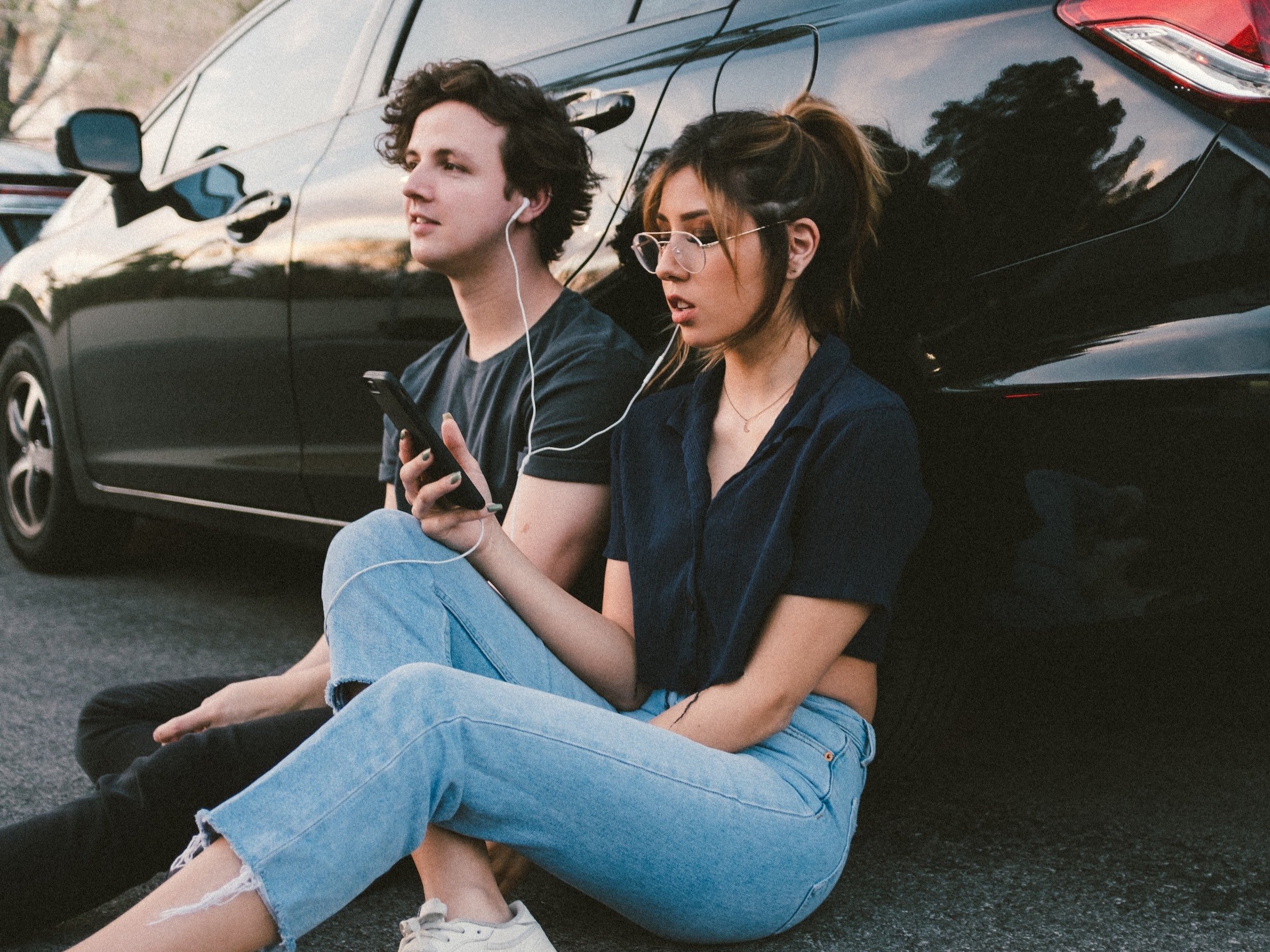 Two friends lean up against a car, share a pair of headphones, and listen to music together.