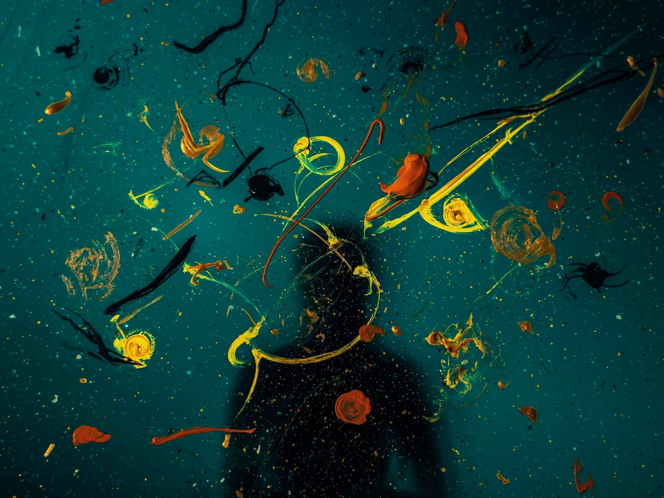 A person's shadow may be seen behind a wall covered in various coloured paint strokes, splodges, and blobs.