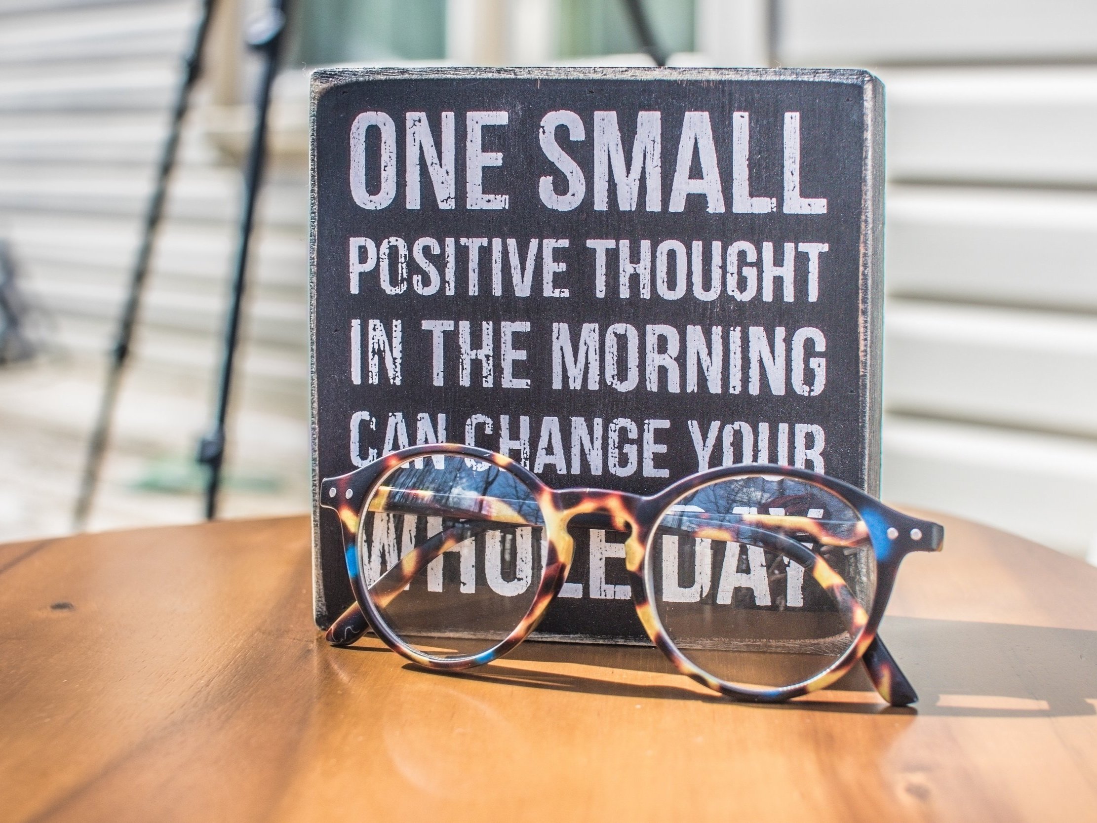 The words "One small positive thought in the morning can change your whole day" are written on a wooden board in front of a pair of sunglasses.