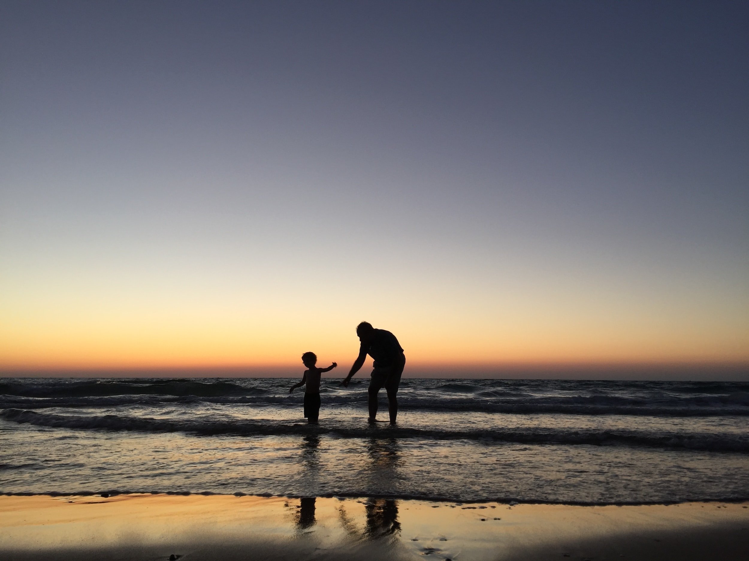 A father and his young son are playing in the sea as the sun sets in the distance.