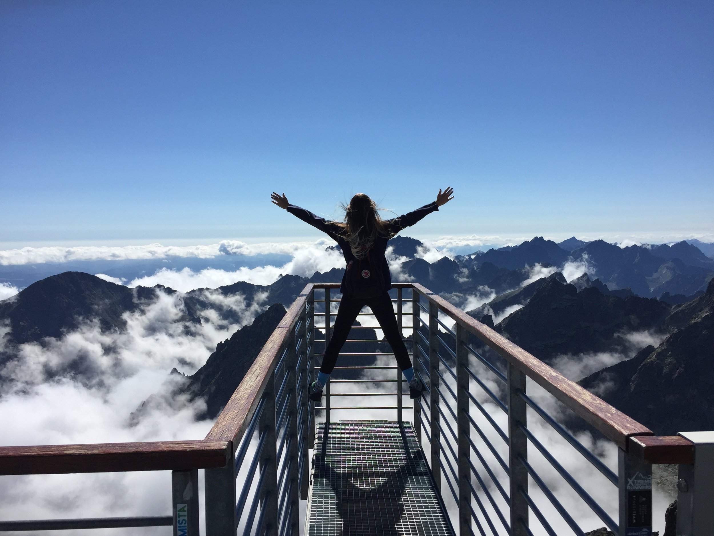 A woman stands on the handrails, arms wide open, facing the mountains and clouds.