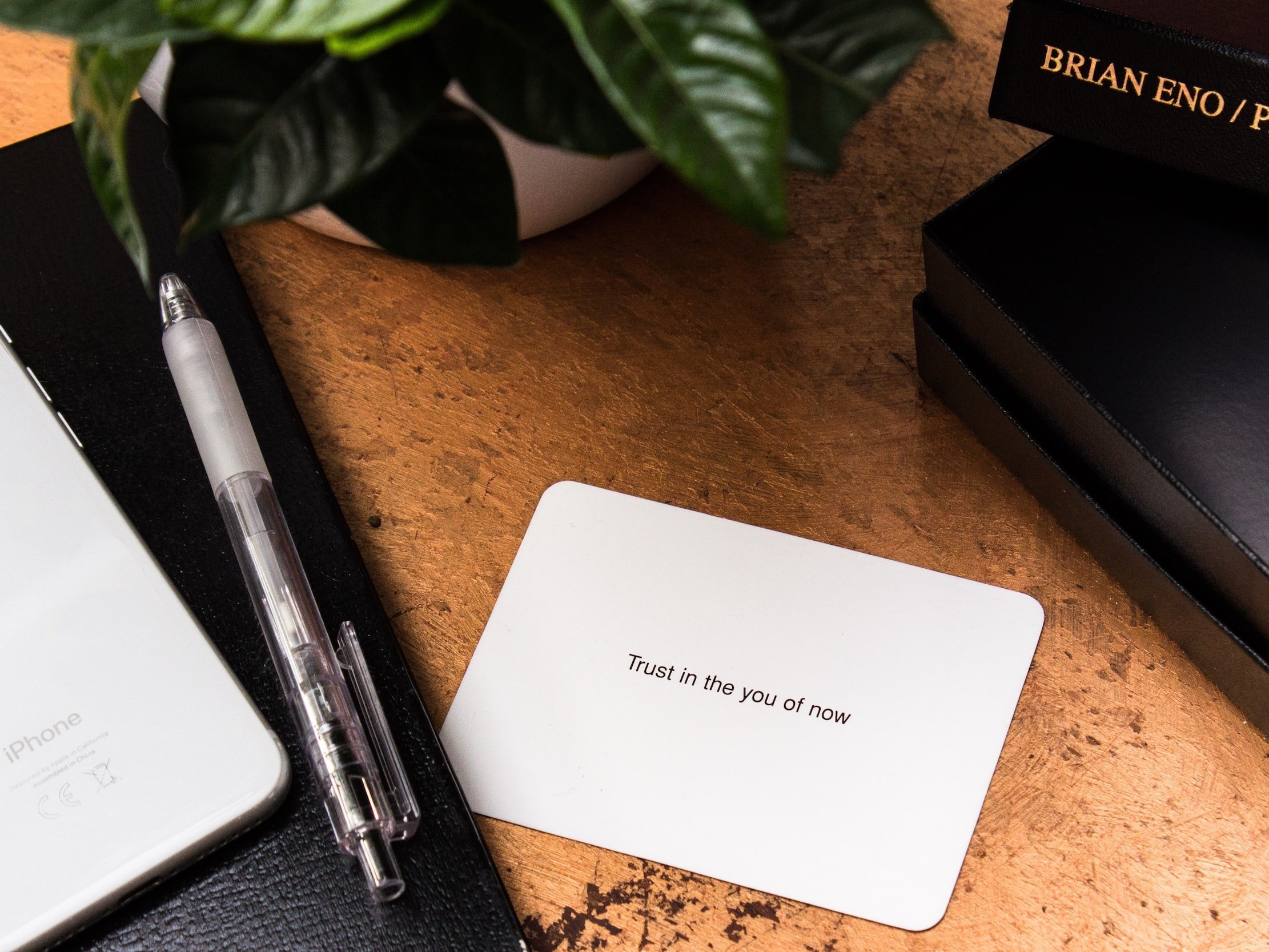 A card with the words "Trust in the You of Now" is displayed alongside an iPhone and a pen.